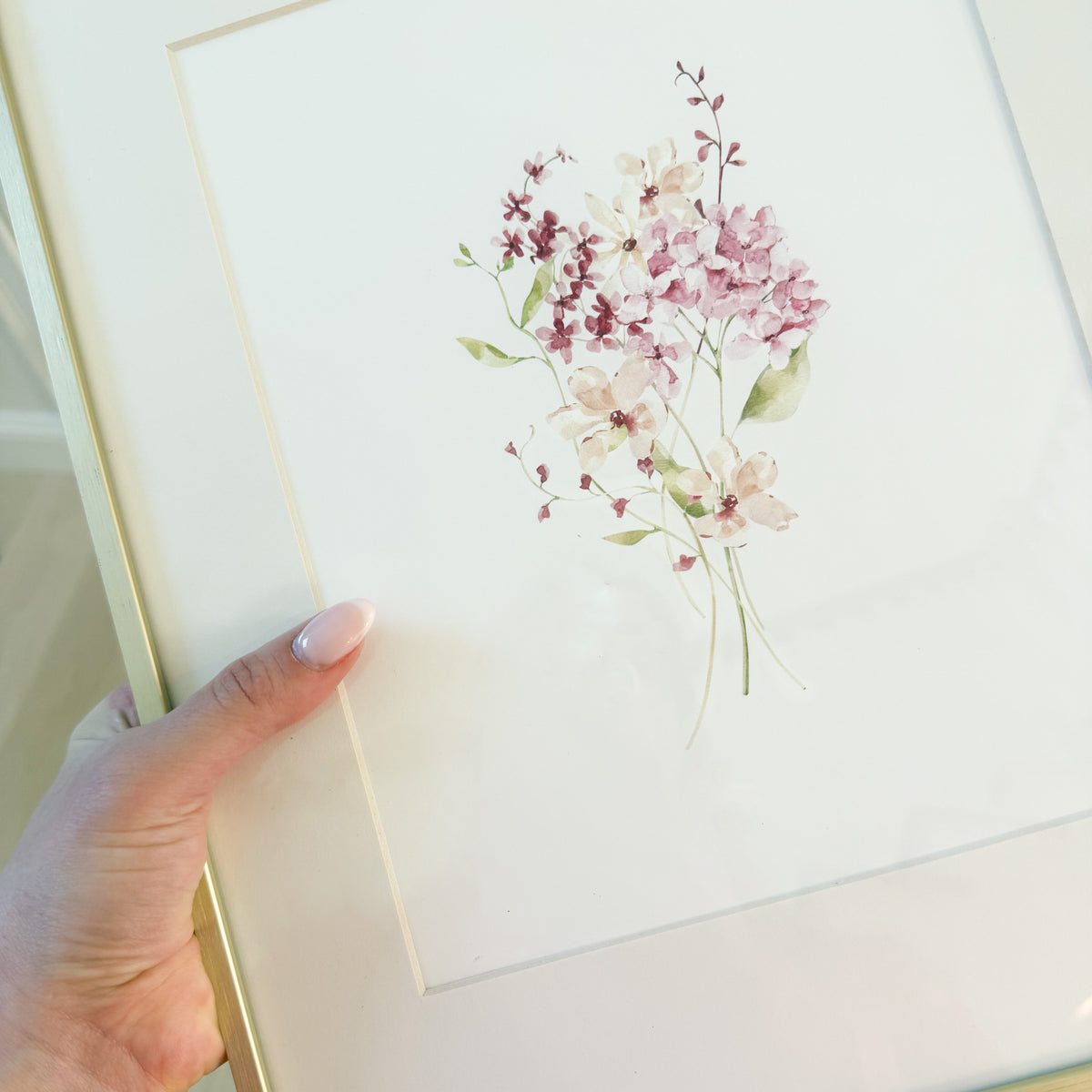 hand holding a gold frame with a picture of a watercolor wildflowers bouquet with pink and burgundy flowers.