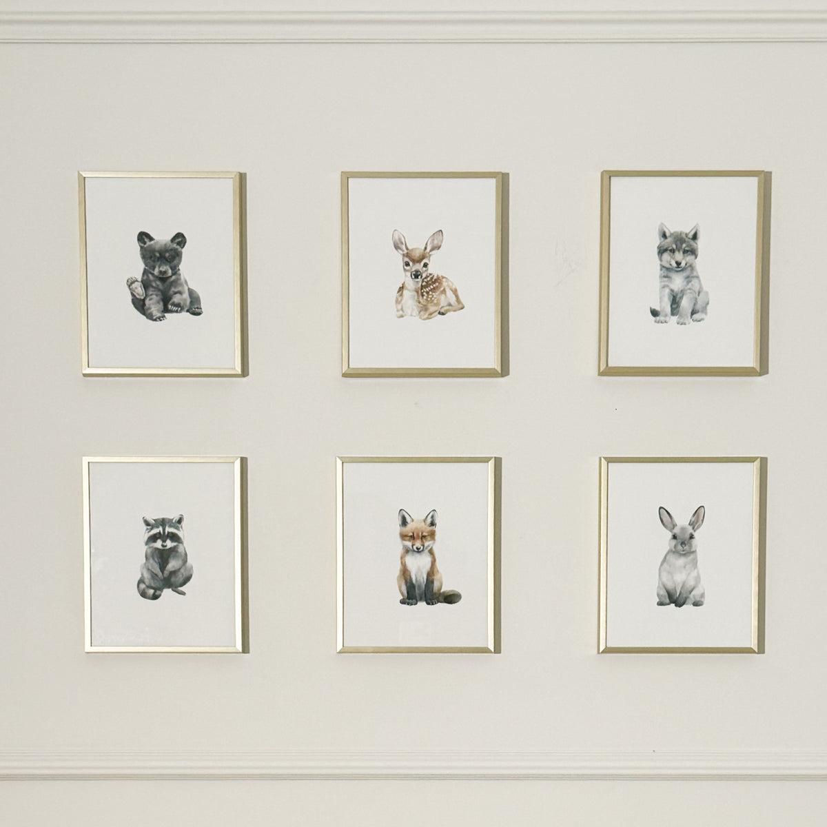 Six framed animal art prints arranged in two rows of three on a light beige wall. The prints feature watercolor-style illustrations of a bear, rabbit, cat, raccoon, fox, and rabbit, each in a thin gold frame.