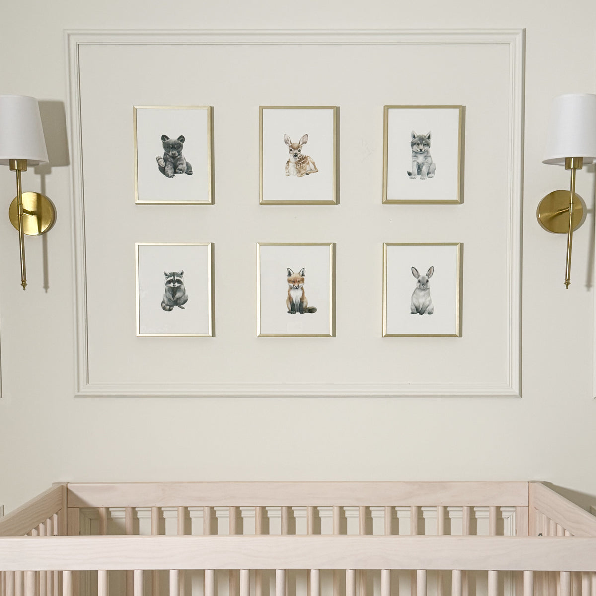 A nursery wall display featuring six framed animal art prints arranged in two columns of three above a light wood crib. The prints show a bear, rabbit, cat, raccoon, fox, and rabbit in soft watercolor style.